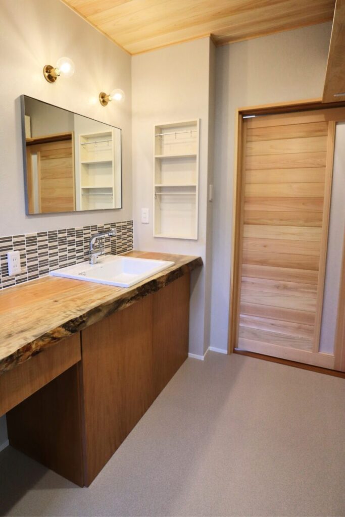 木の天井とカウンターが広がる洗面・ランドリースペース。洗濯機や収納棚が整い、タイルの壁と照明が明るく落ち着いた雰囲気をつくる。