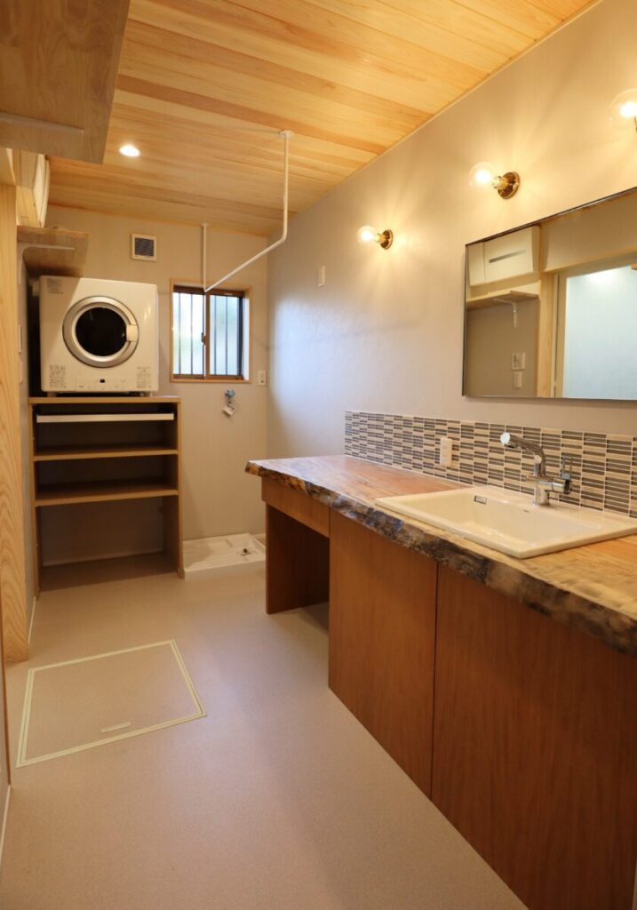 木の天井とカウンターが広がる洗面・ランドリースペース。洗濯機や収納棚が整い、タイルの壁と照明が明るく落ち着いた雰囲気をつくる。
