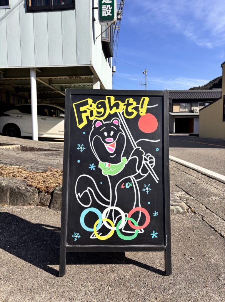 ブラックボードにミラノｺﾙﾃｨﾅオリンピックのマスコットキャラクターのティナ（ミロ）を書いて日本の国旗を書いてファイトと応援している