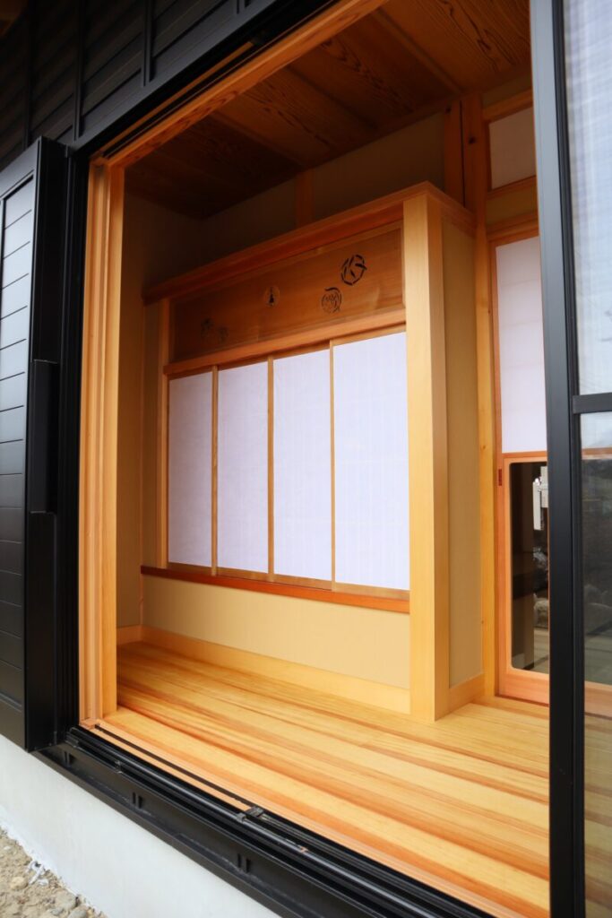 黒い外壁に大開口のガラス戸が設けられ、内部には木の温もりが広がる和室が見える外観。欄間と障子が美しく納まった、和と現代が調和する佇まい。