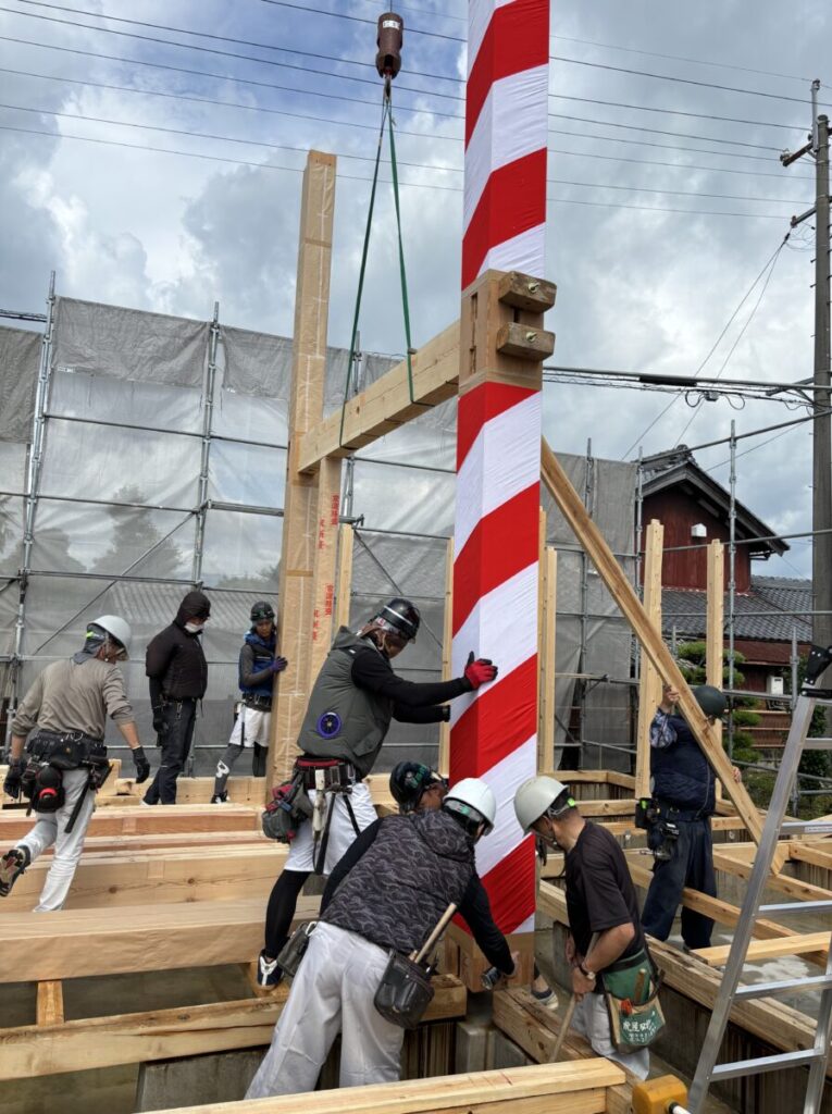 木造住宅の建て方作業の様子。職人たちが柱や梁を組み上げ、紅白の布を巻いた大黒柱が立つ中で工事が進む。