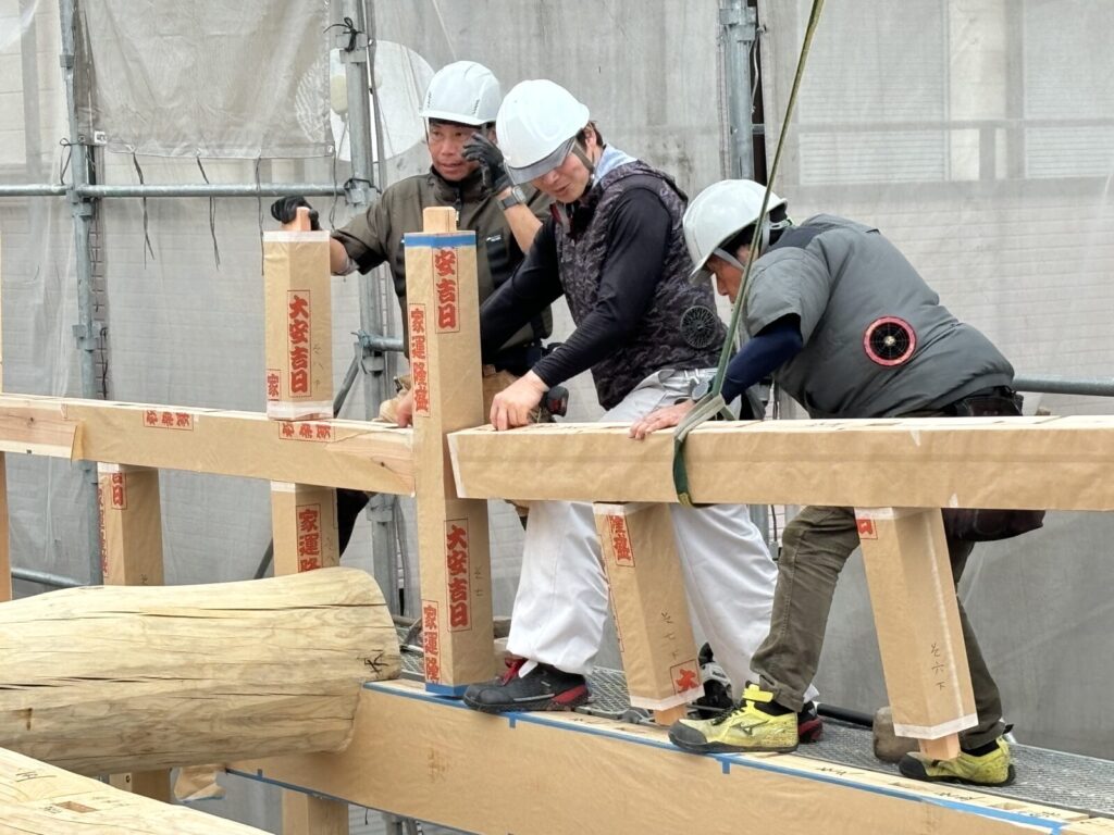 木造住宅の建て方作業の様子。職人たちが柱と梁を組み上げ、紅白の巻き柱が立つ中で丁寧に木組みを進めている。