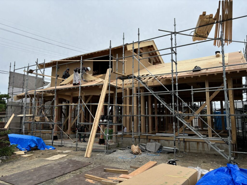 足場が組まれた木造住宅の建築現場。瓦屋根の形が見える骨組みが立ち上がり、庭の松が佇む中で工事が進む様子。