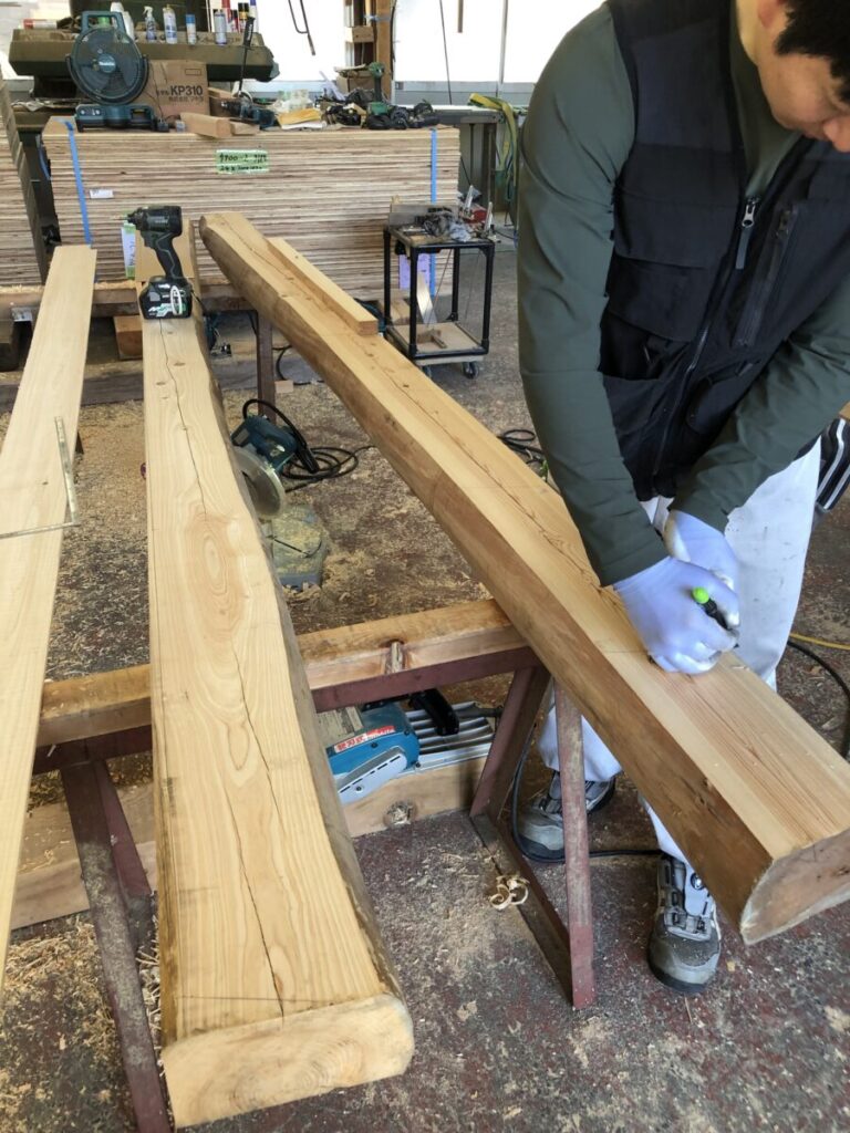 作業場で職人が木材に墨付けを行っている様子。大きな木材に丁寧に印を付け、周囲には工具や材料が並ぶ。