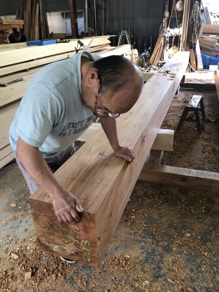 作業場で職人が木材に墨付けを行っている様子。大きな木材に丁寧に印を付け、周囲には工具や材料が並ぶ。