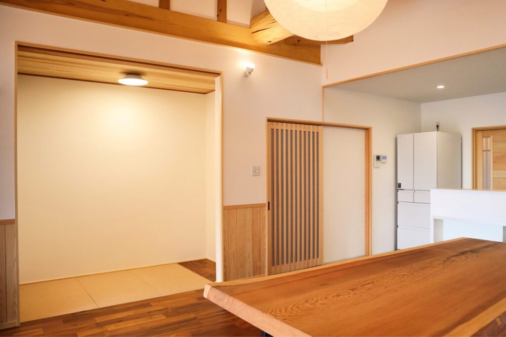 木の梁と板張り天井が広がる和モダンの室内。畳スペースと木製建具が調和し、無垢材のテーブルが置かれた温かみのある空間。