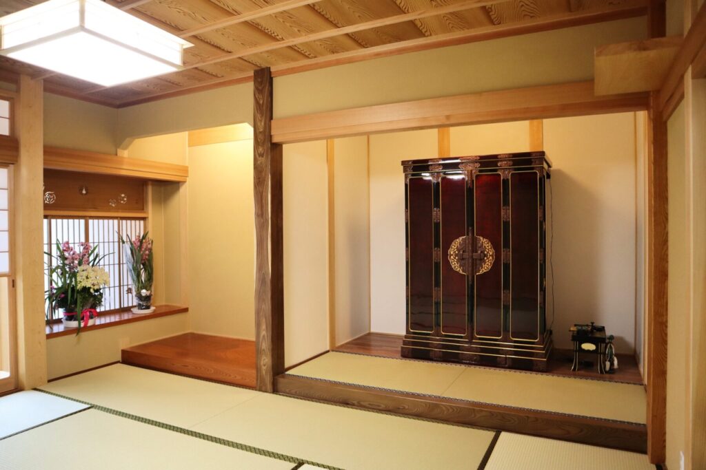 畳と障子が広がる和室。床の間に花が飾られ、格子天井と木の建具、仏壇が静かな佇まいをつくる。